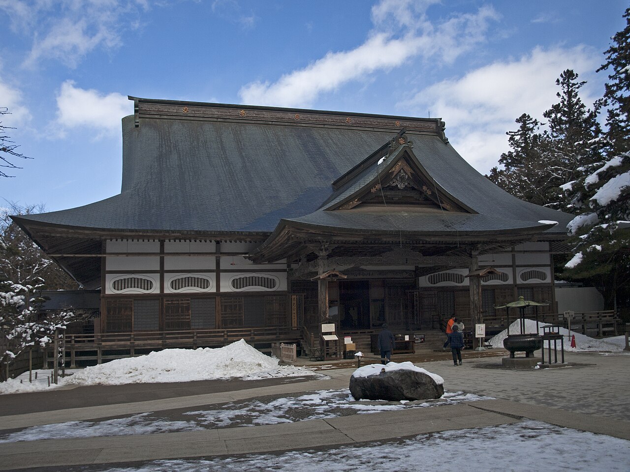 Front of a large wooden temple hall with a wide dark sloping roof and white walls with oval lattice windows, snow on the ground and a few visitors near the entrance.