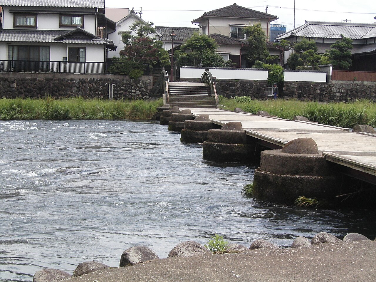 A stepping-stone submersible bridge crossing the fast-flowing Mikuma River in Hita with houses on the far bank