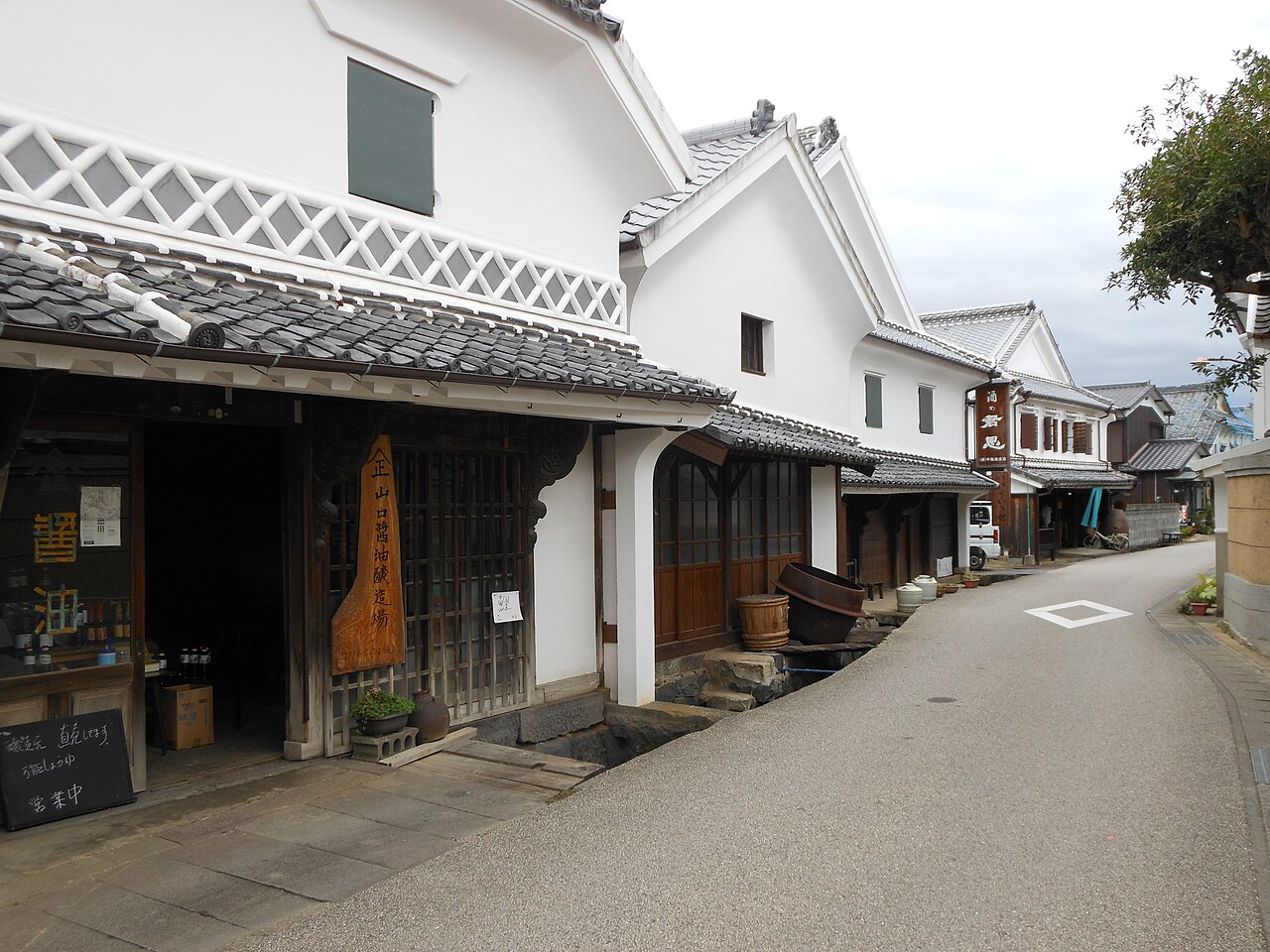 A quiet paved street runs past white-plastered storehouses with gabled tile roofs and latticed eaves, with wooden barrels and a large clay vat outside a shop entrance on the left and a wooden hanging sign further along.