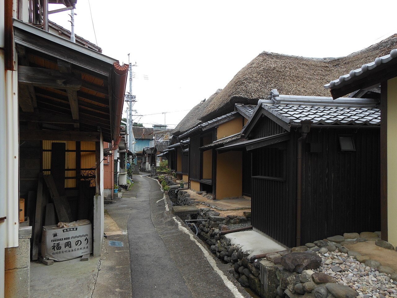A narrow paved lane runs between dark timber houses with ochre plaster walls and a large thatched roof, with a low stone-lined channel along the right side and tiled-roof houses stretching ahead.