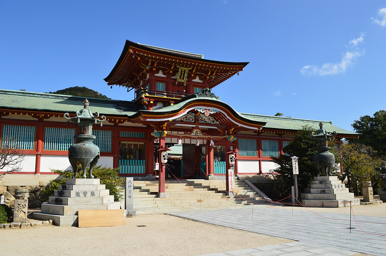 The vermilion two-storey Romon gate of Hofu Tenmangu above a wide stone stairway, flanked by bronze lanterns under a clear blue sky