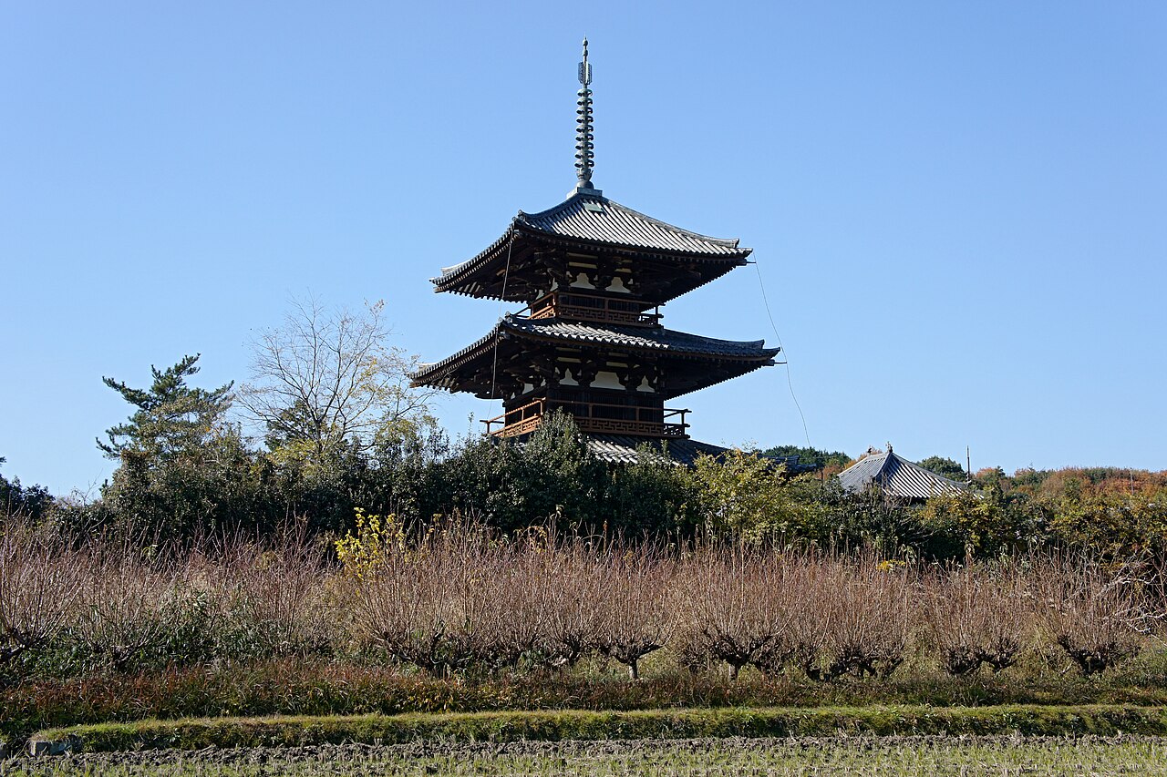 Hokiji's three-storey pagoda standing behind a strip of dry winter fields and bare shrubs, with low wooded hills and a temple roof beside it