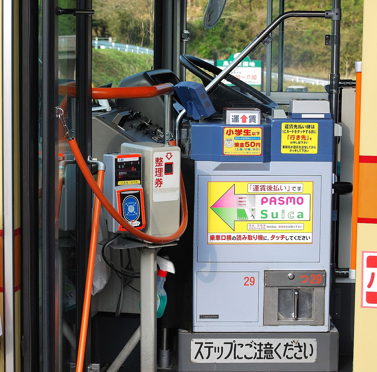 Driver's area of a Japanese bus seen from the door: numbered-ticket dispenser and an IC card touch reader on a pole, next to a fare box with PASMO and Suica instructions
