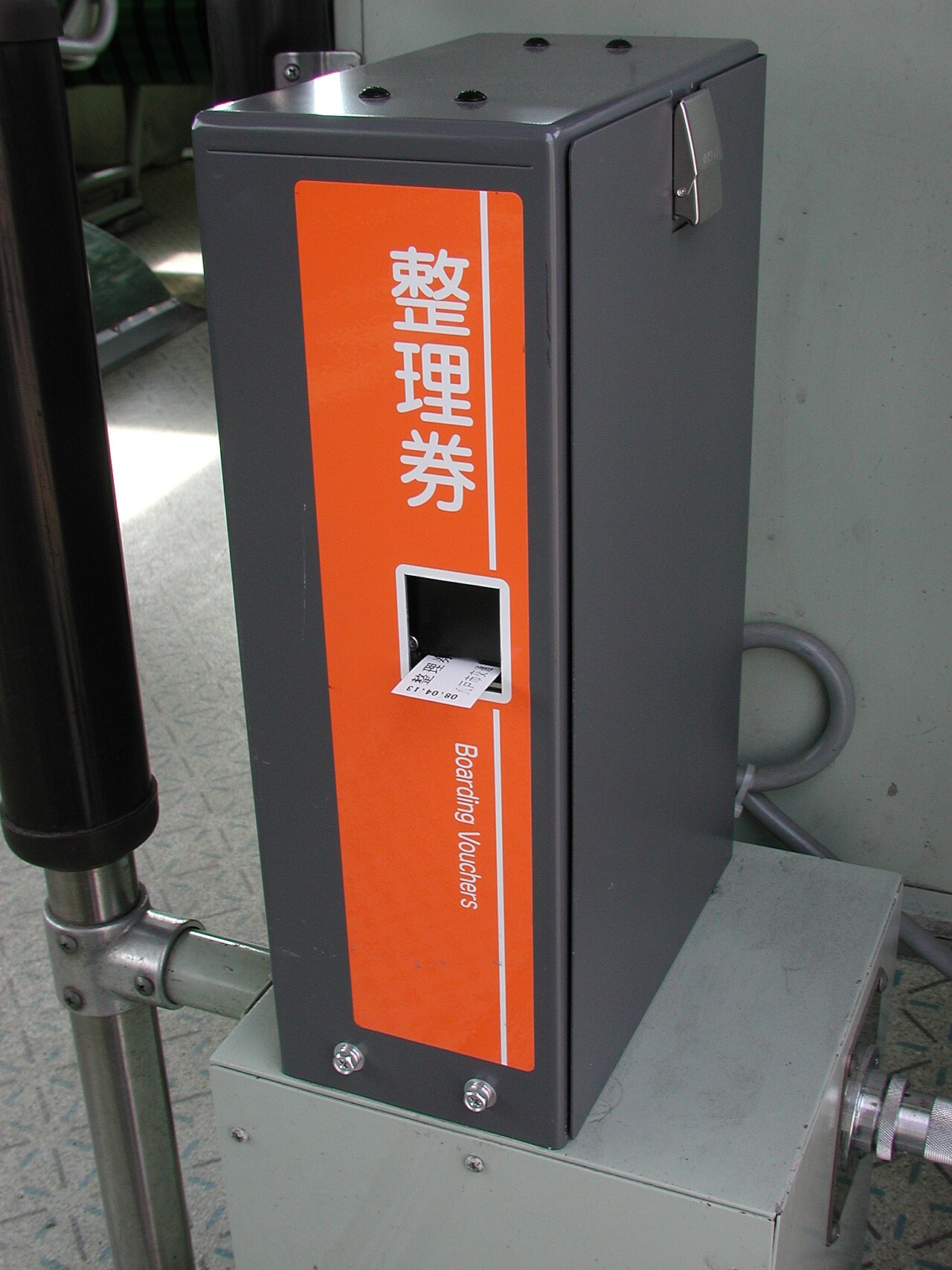 Orange boxed numbered-ticket dispenser labeled 整理券 / Boarding Vouchers inside a Japanese local bus, with a small printed ticket sticking out of the slot