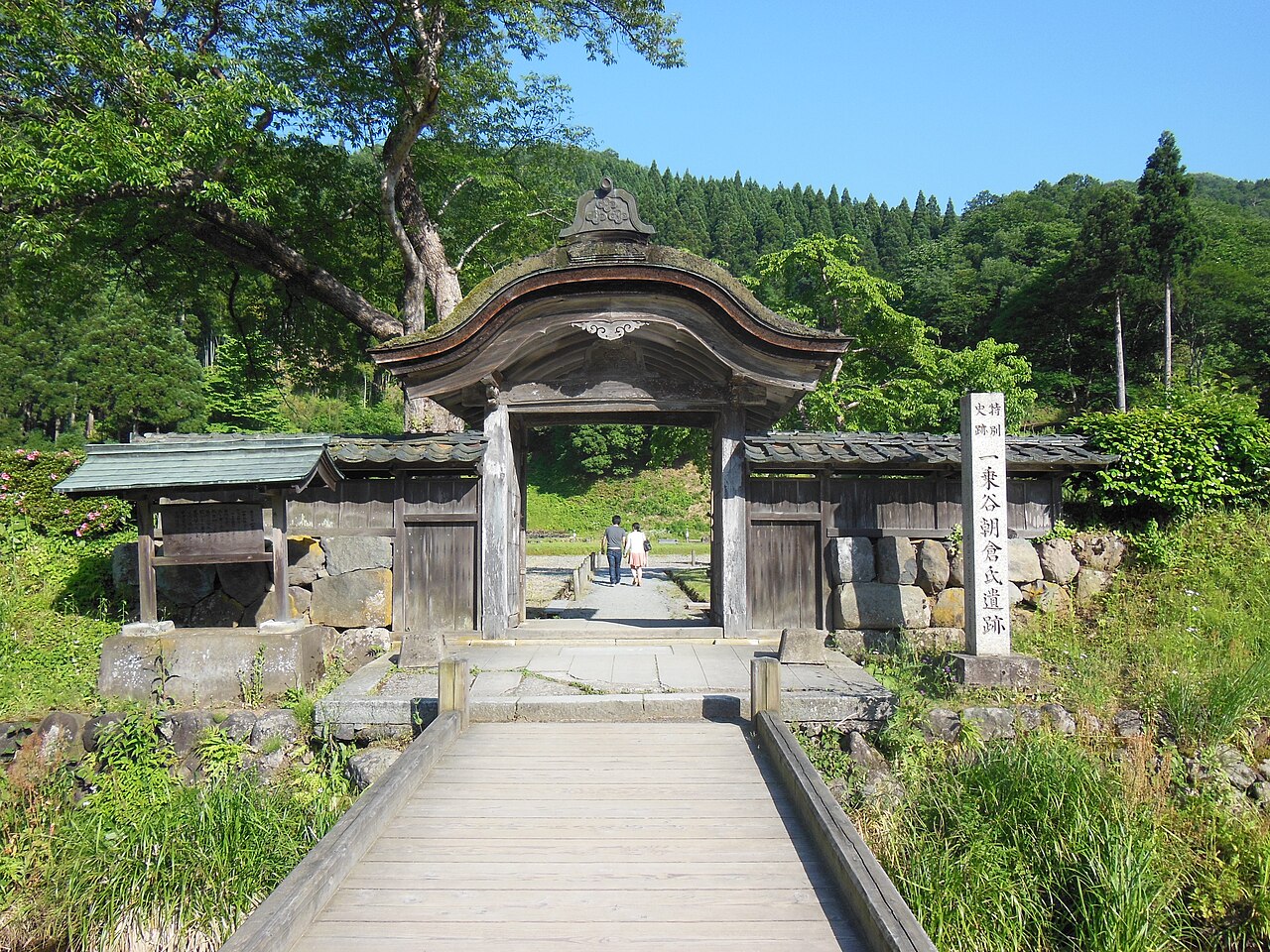 The Karamon gate at the Asakura residence site in Ichijodani, a curved-roof gate reached by a plank footbridge, with a stone marker beside it and cedar slopes behind