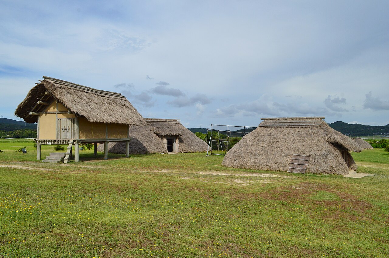 Reconstructed thatched-roof raised storehouse and pit dwellings on the grassy Harunotsuji archaeological site on Iki Island.