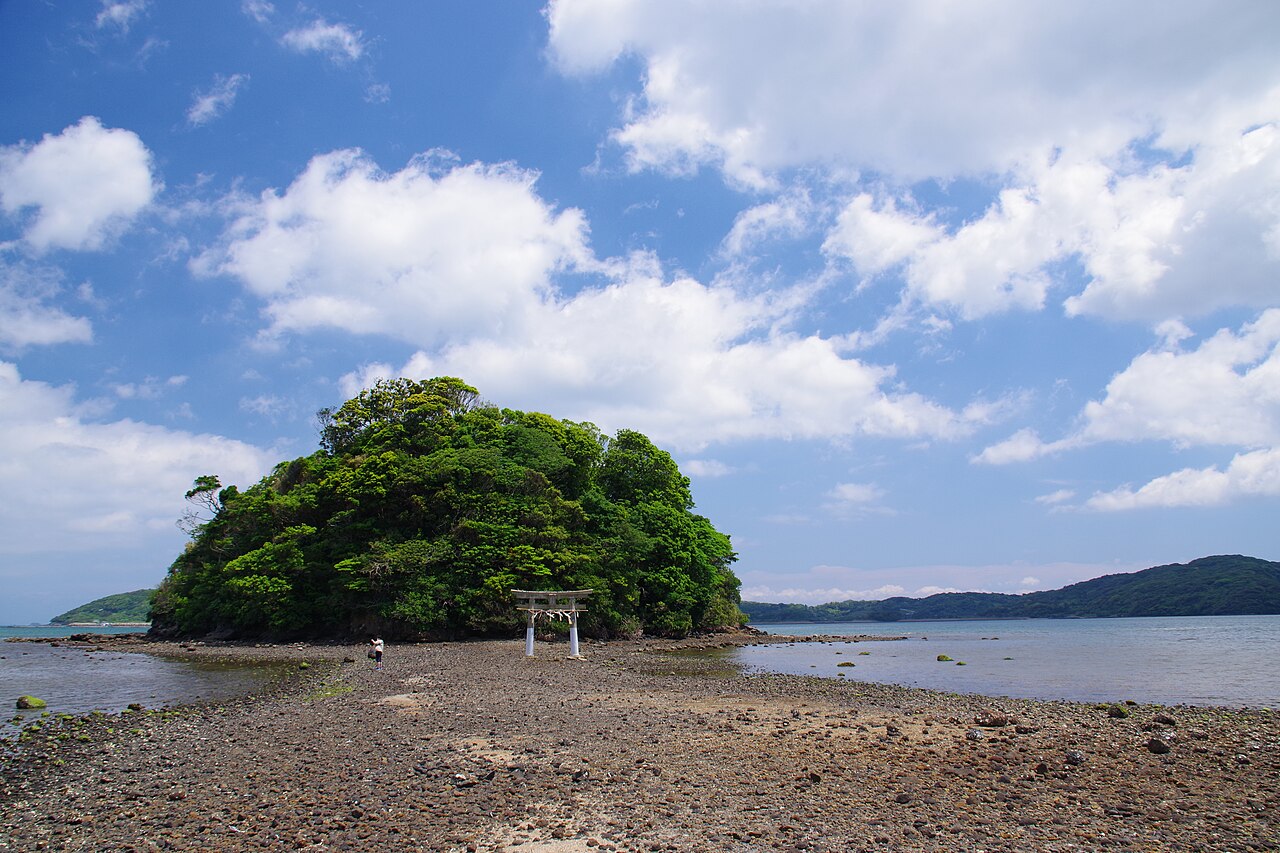 A small wooded islet with a white torii gate at its foot, reached across exposed tidal flats, under a blue sky (Kojima Shrine, Iki Island).