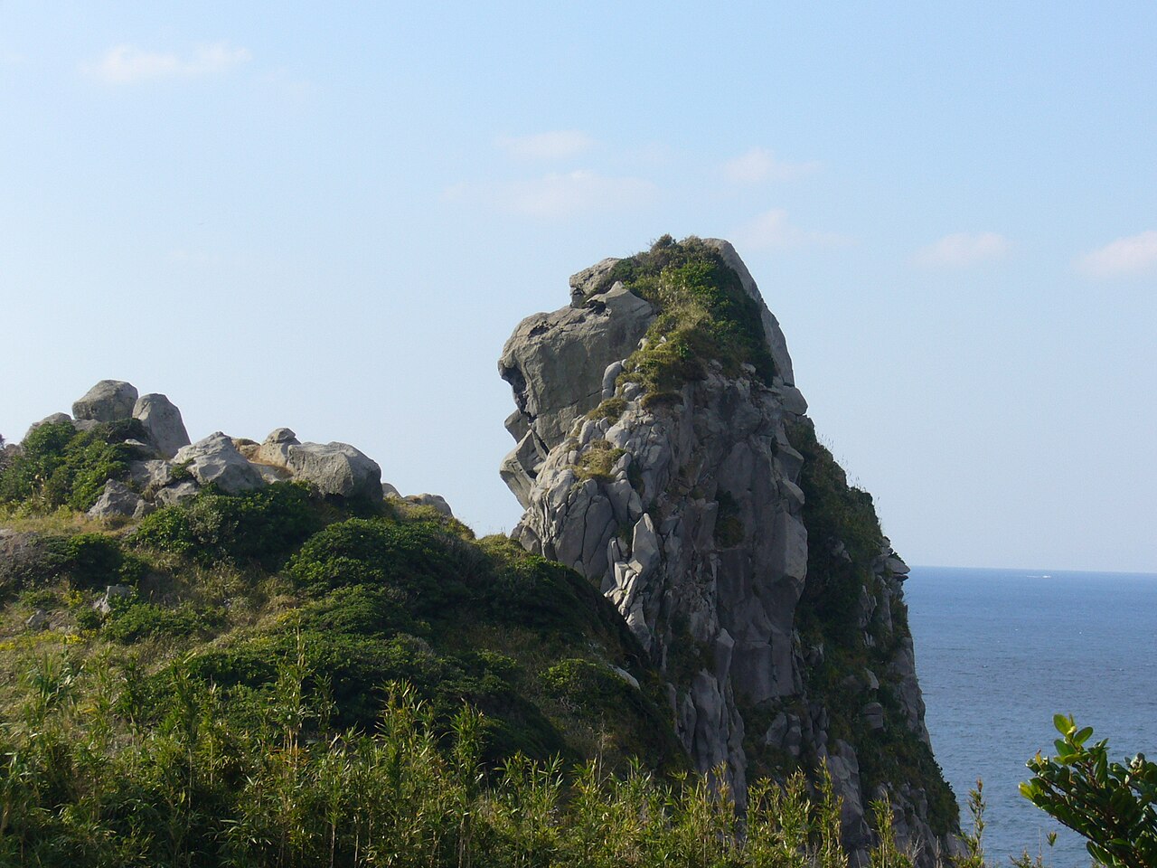 A tall weathered rock outcrop on a green Iki Island headland, its profile resembling an ape's face, with the sea beyond.