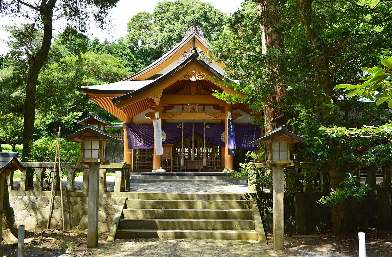 Front view of Sumiyoshi Shrine's worship hall on Iki Island, with stone steps, lanterns and a purple curtain among tall trees.