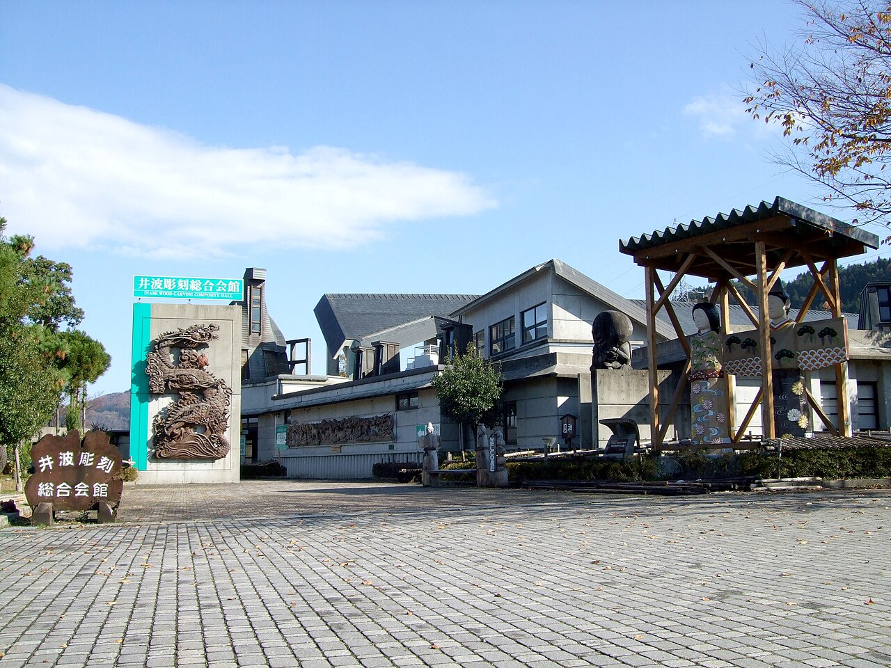 Low buildings with dark tiled roofs behind a stone-paved plaza in Inami, with a tall carved wooden figure on a pillar at the left under a blue sky