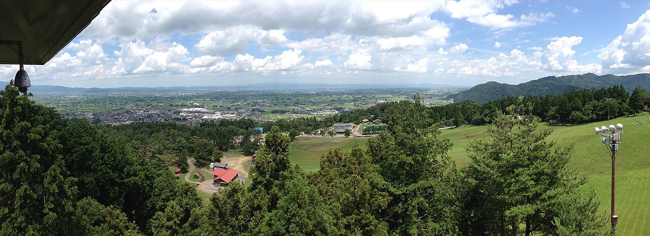 A wide view over a green valley and plain with farmland and scattered buildings under a cloudy blue sky, seen from a wooden lookout roof