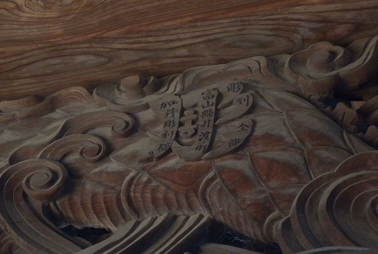 A close view of deeply carved dark wood relief of swirling waves with a carved inscription, on the front of a shrine hall