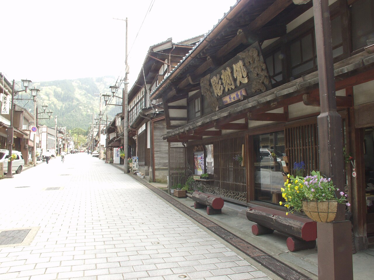 A paved street lined with dark wooden merchant houses and a large carved wooden shop sign on the right, with green mountains at the far end