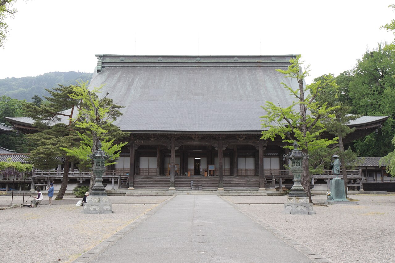 A broad wooden temple main hall with a vast grey tiled roof seen along a paved approach, with pine trees at both sides and mountains behind