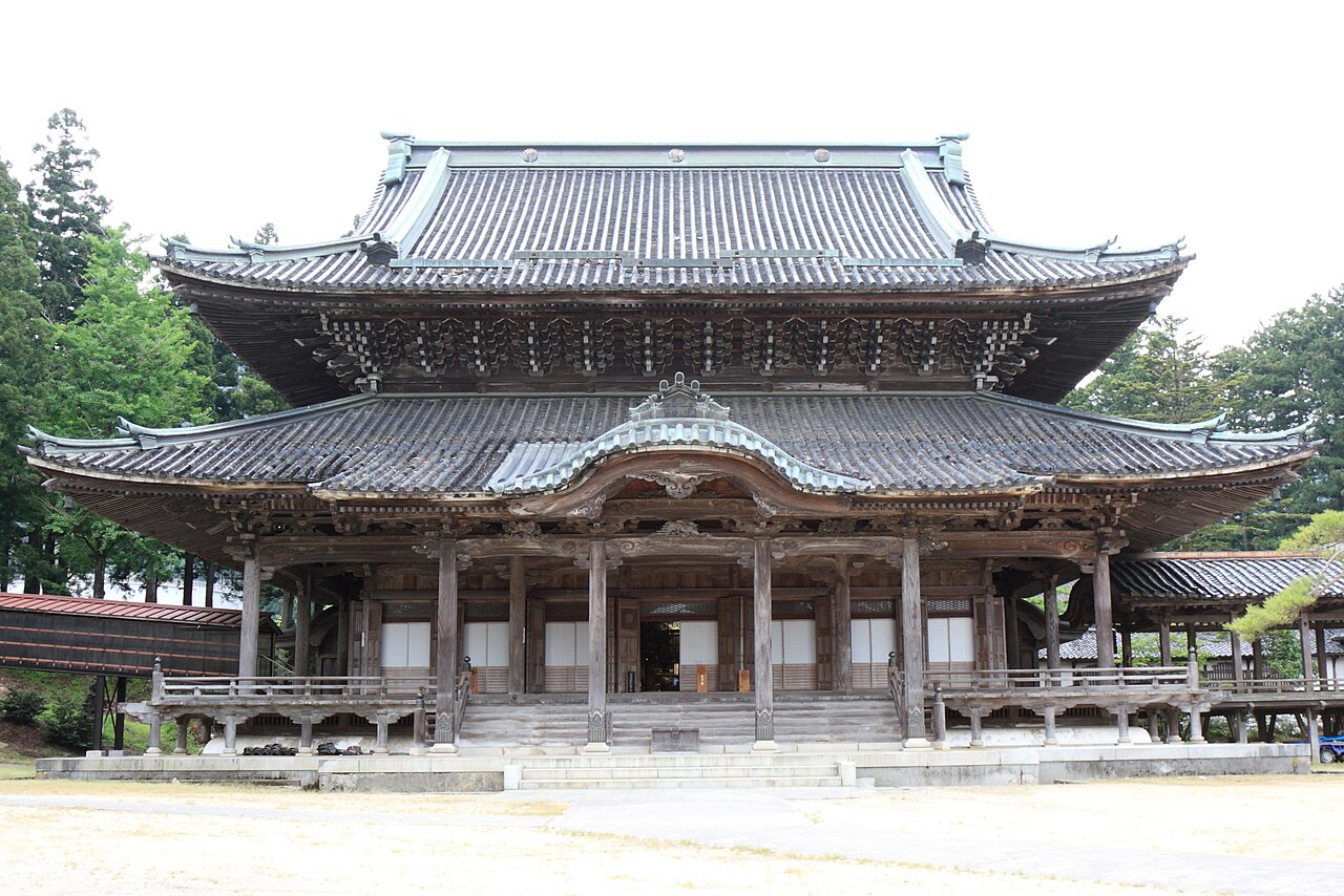 A two-storey wooden temple hall with a layered tiled roof and carved eaves standing at the end of a wide gravel courtyard, with forested hills behind