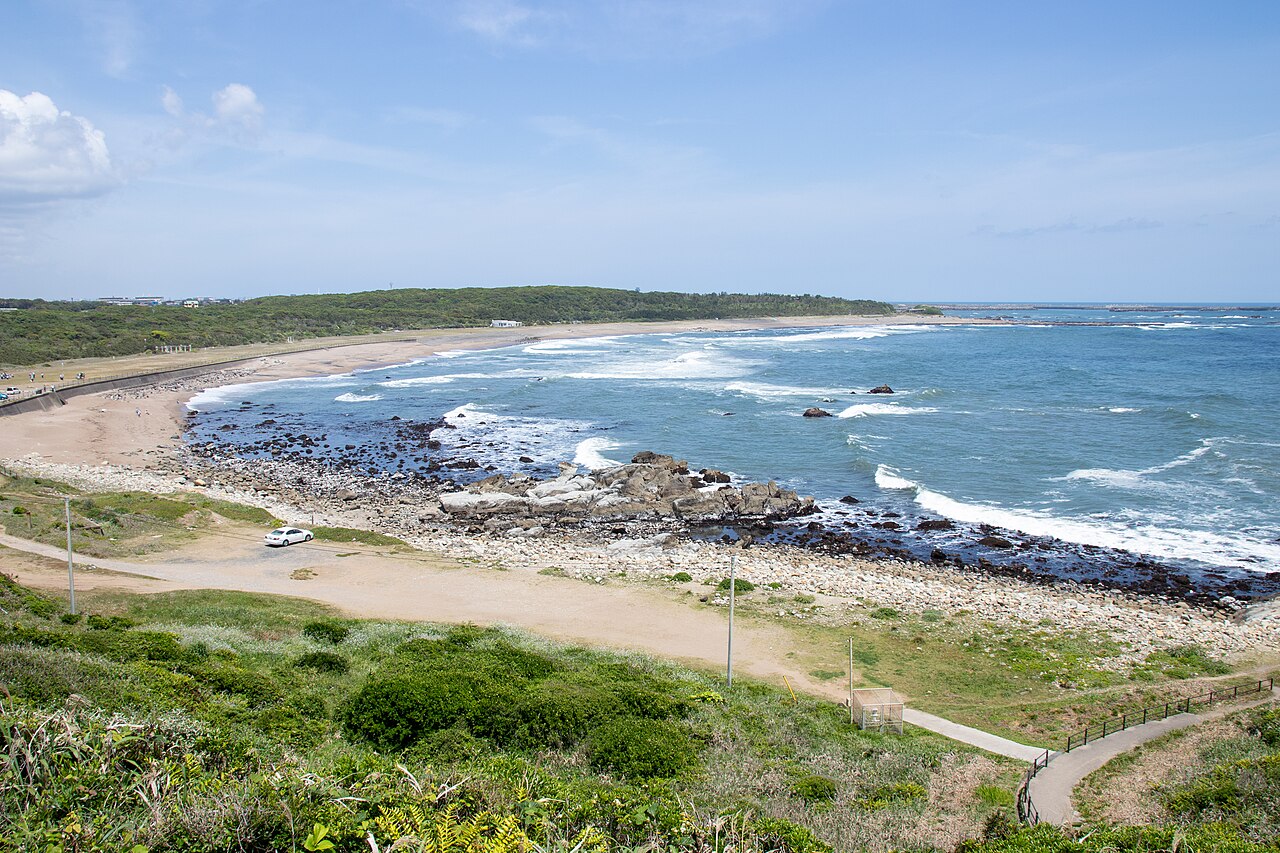 A curving sandy bay and surf-washed rocks along the coast at Cape Inubo, seen from the grassy slope above