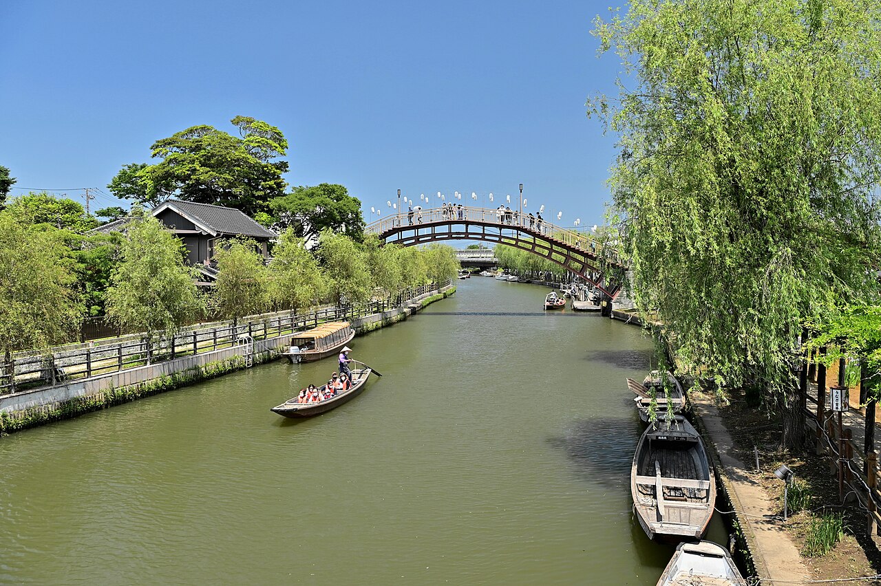 A narrow boat carrying passengers in orange life jackets moves along a wide green canal toward an arched wooden bridge strung with paper lanterns, willows hanging over the banks under a clear blue sky