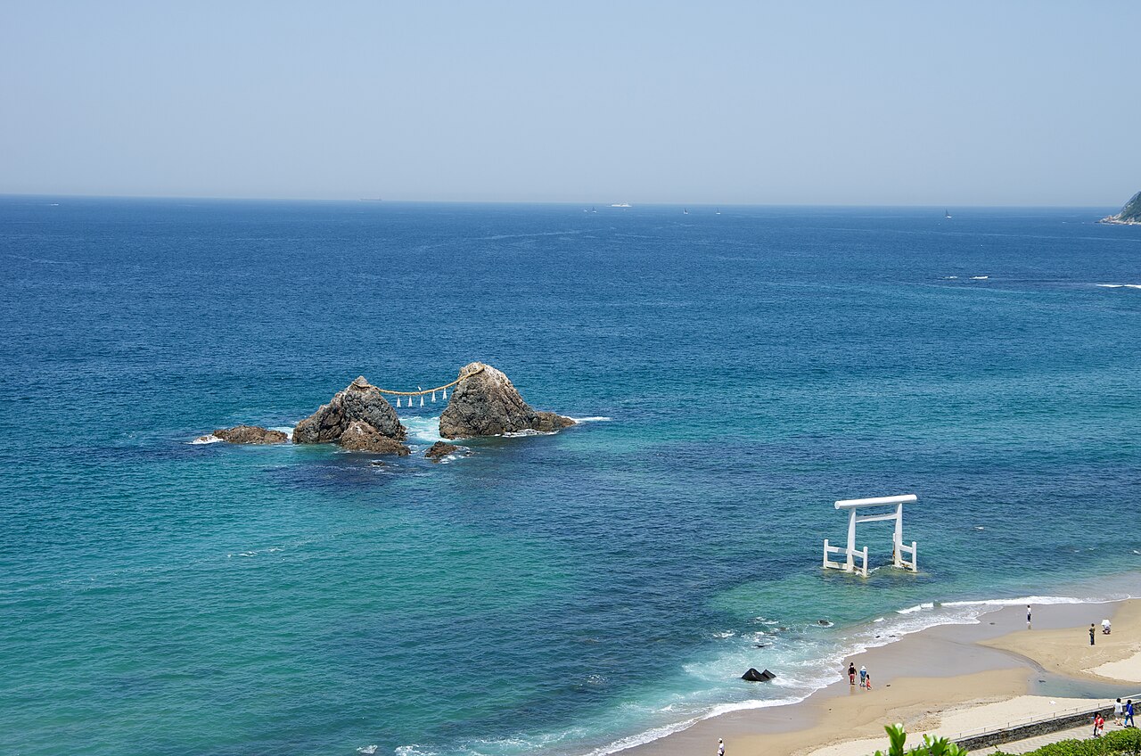 The white torii standing in the shallows off Sakurai Futamigaura beach with the paired Meoto Iwa rocks beyond, seen from above the clear blue sea