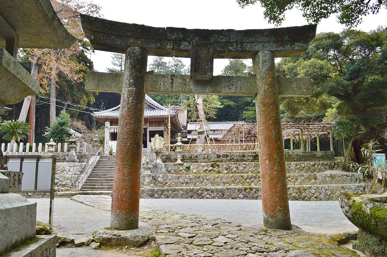 A weathered stone torii at Takasu Shrine in Itoshima with stone steps and a worship hall visible behind it