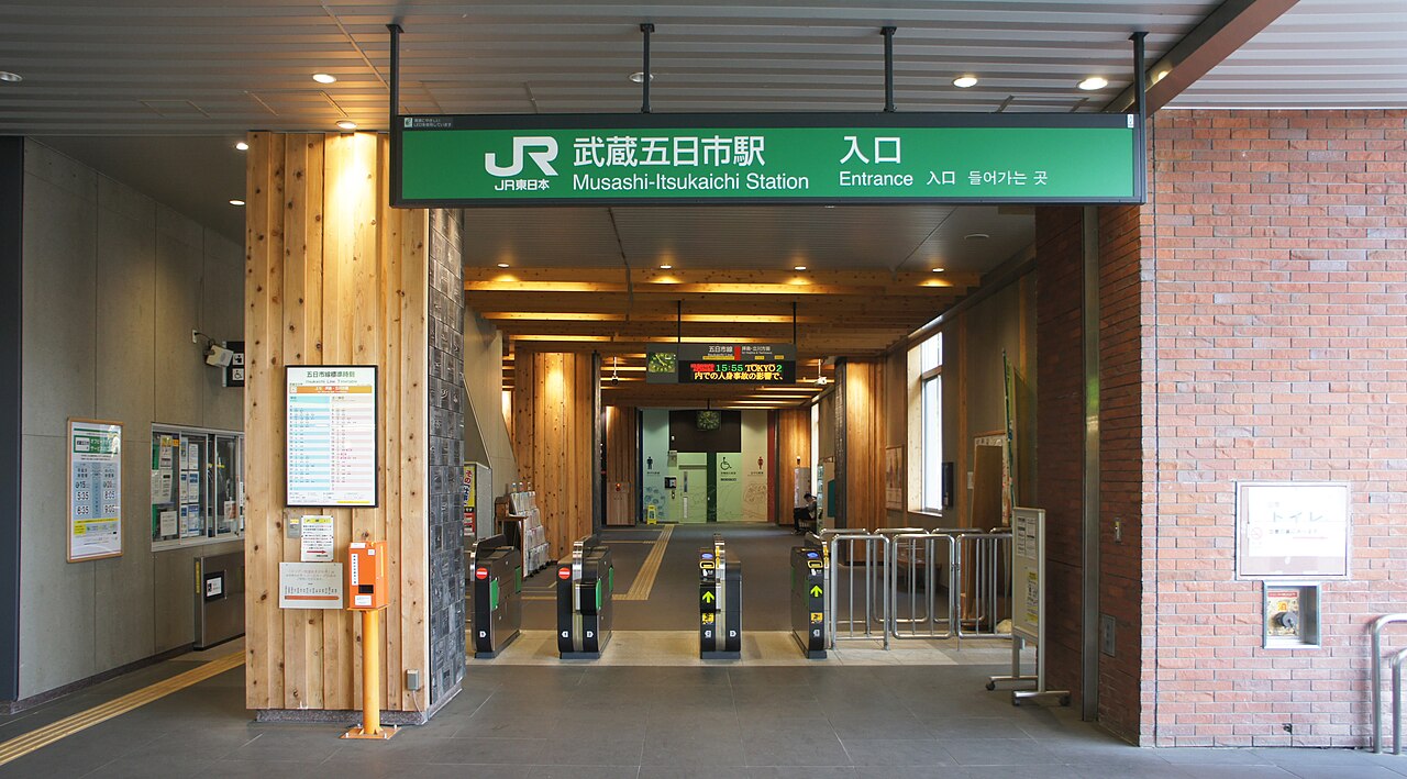 The ticket gates of Musashi-Itsukaichi Station under a green bilingual sign, with warm wooden pillars and a paper timetable posted at the entrance.
