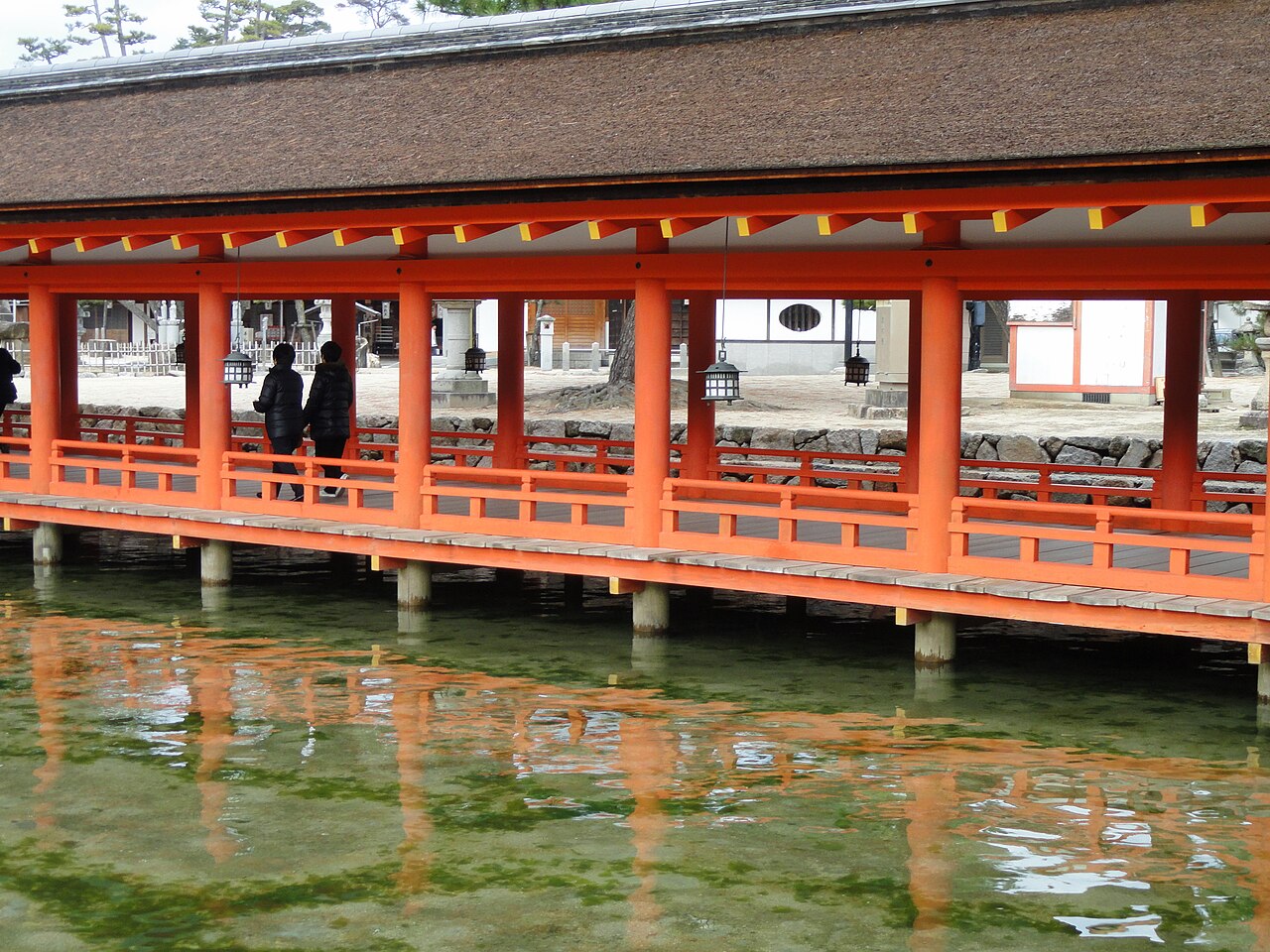 A long vermilion covered corridor of Itsukushima Shrine raised on posts above still, green-tinted water, with its reflection below and two small figures inside.