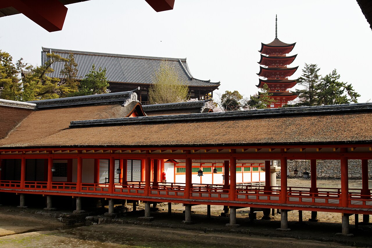 The red five-storied pagoda rising behind a dark-roofed building, with a vermilion shrine corridor on stilts in the foreground and a pale sky above.