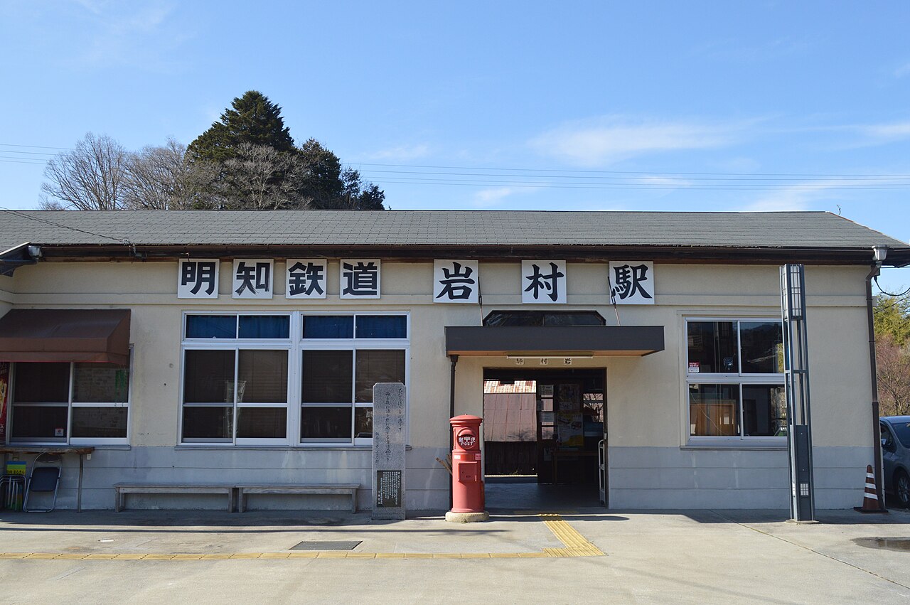 The low station building with large kanji signs reading Akechi Railway Iwamura Station, a red cylindrical post box and a stone marker out front.