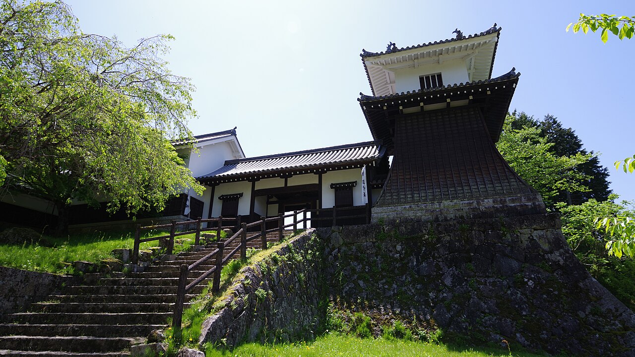 A reconstructed gatehouse and drum tower standing above a stone-walled slope and a flight of stone steps, with fresh green trees and a blue sky.