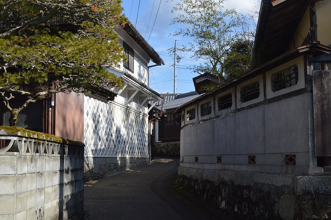 A narrow lane between a white diamond-patterned namako-style wall and a plastered wall with small barred windows above a stone foundation.