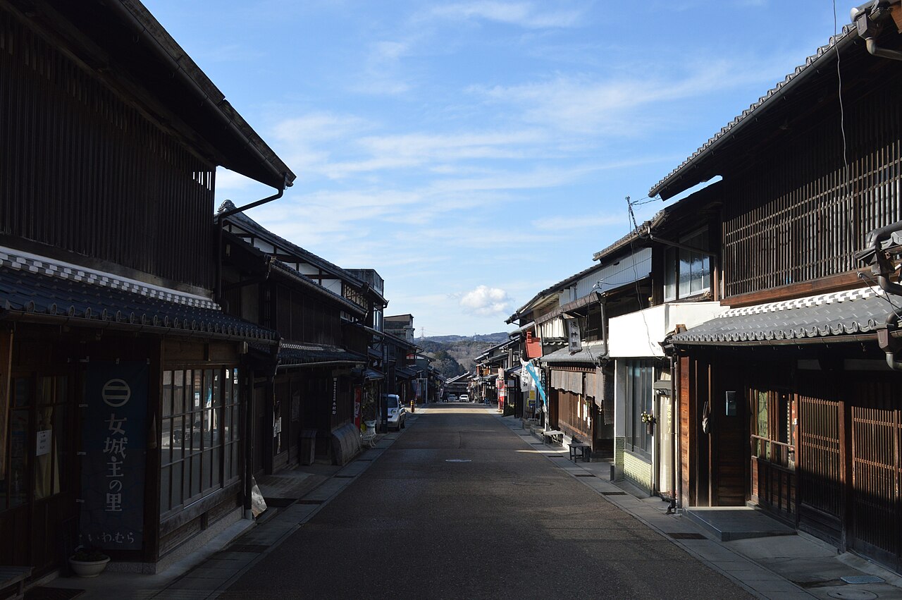 A street lined with dark wooden townhouses, a blue noren curtain reading Onna-jo-shu no Sato at left and low hills in the distance.
