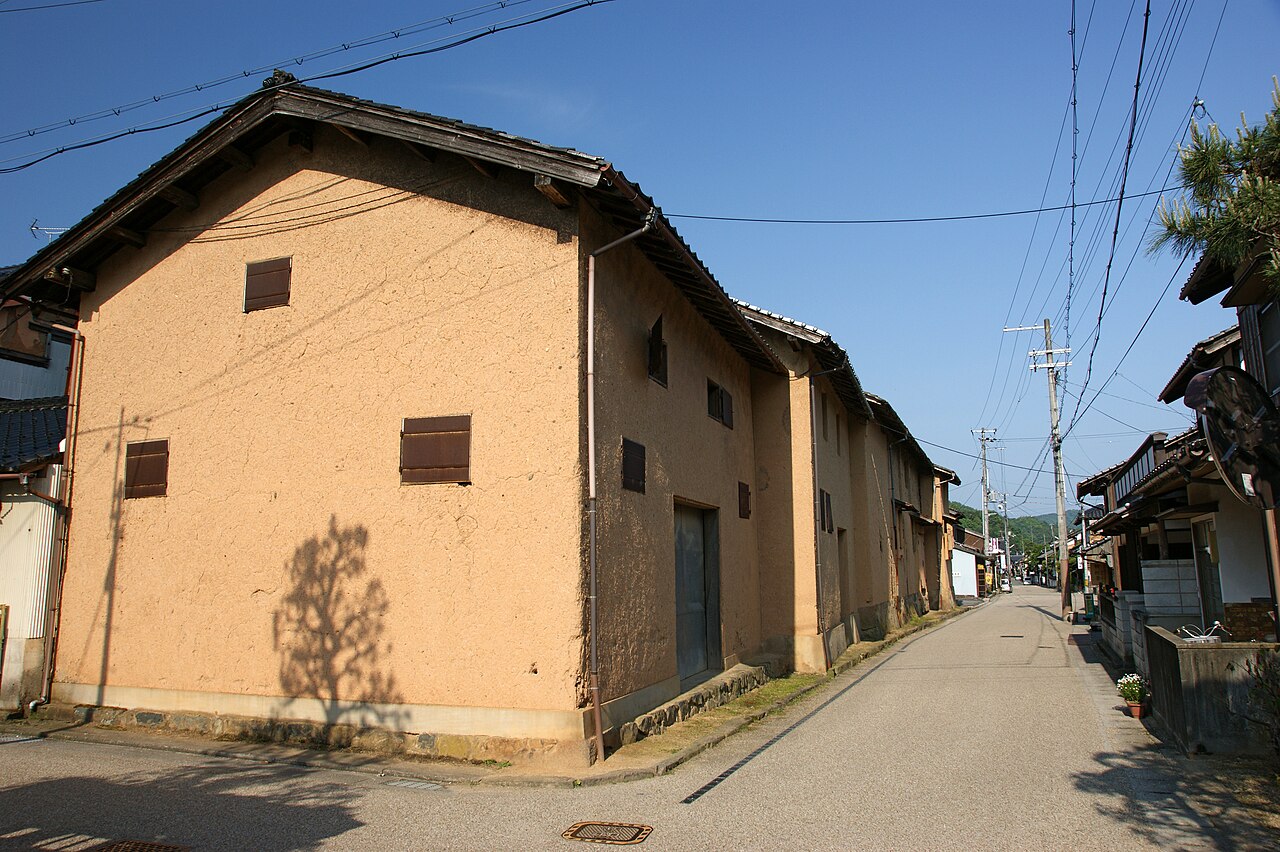 A long earthen-walled storehouse with small shuttered windows on a sunny corner of a quiet Izushi lane, with utility poles and a narrow road running into the distance