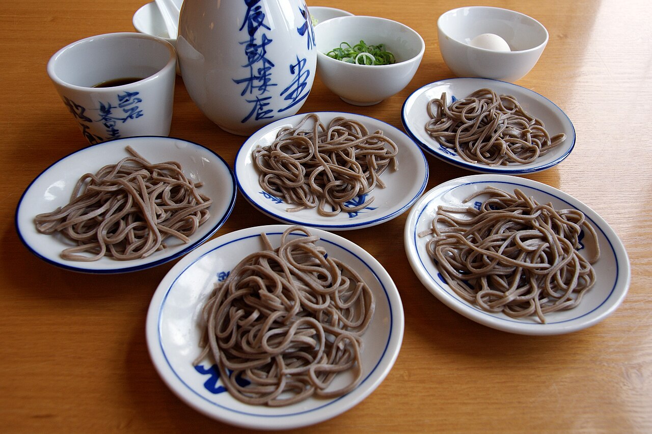 Five small white-and-blue plates of Izushi's dish soba, each holding a bundle of buckwheat noodles, with a dipping cup, grated garnish, chopped scallion and a raw egg