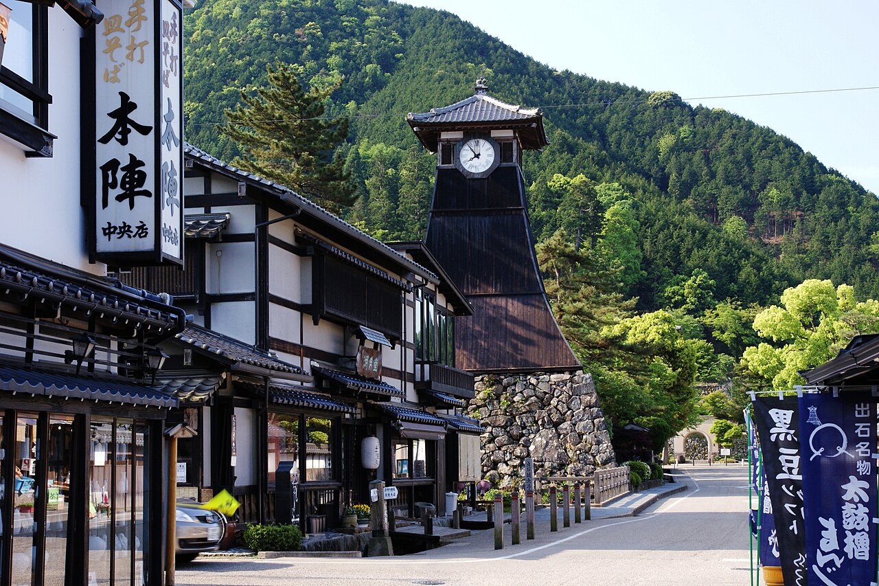 Shinkoro, the dark wooden clock tower of Izushi, rising from a stone base at the end of a quiet street of soba shops and hanging signs, with a green wooded hill behind