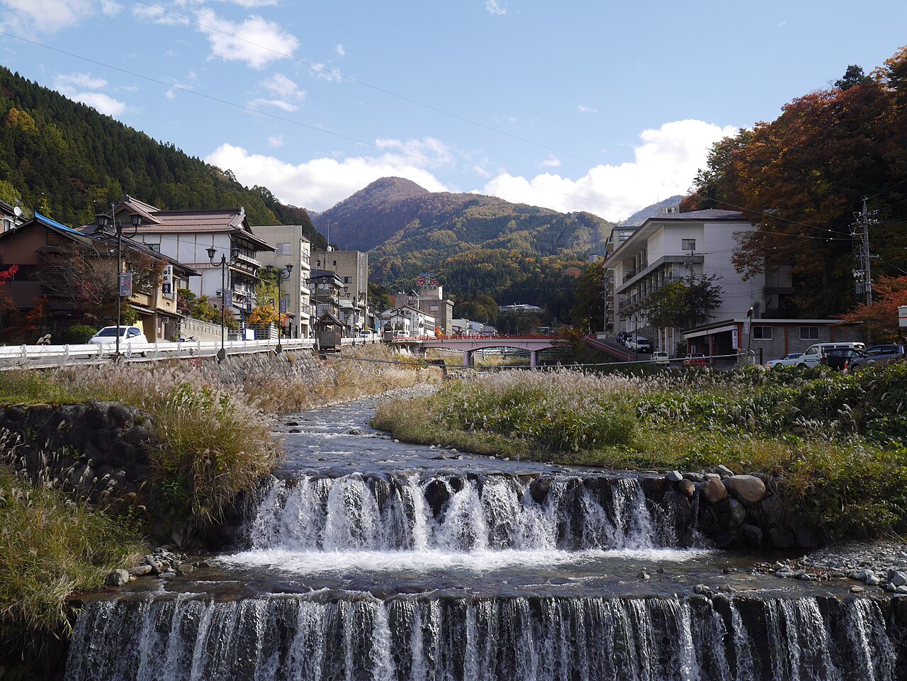 Ryokan buildings line a stream with a low cascade in an old hot-spring town, with mountains behind.