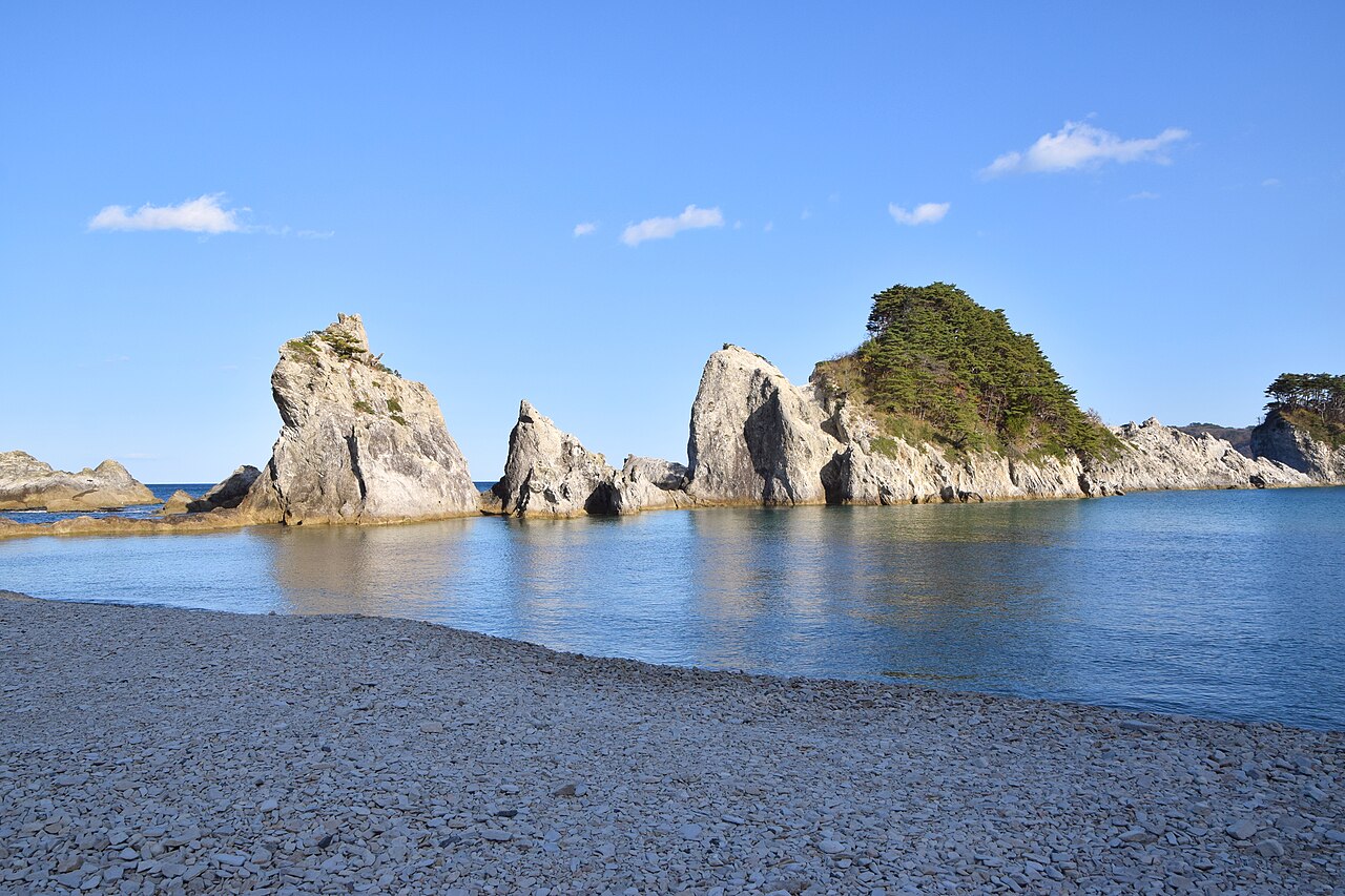 Pale jagged rock stacks topped with pine trees rising from calm water beyond a beach of grey pebbles