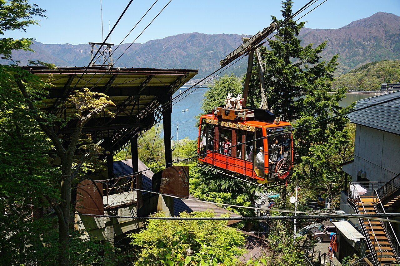 A red Kachikachiyama Ropeway car climbing through green trees, with Lake Kawaguchi and mountains behind it.