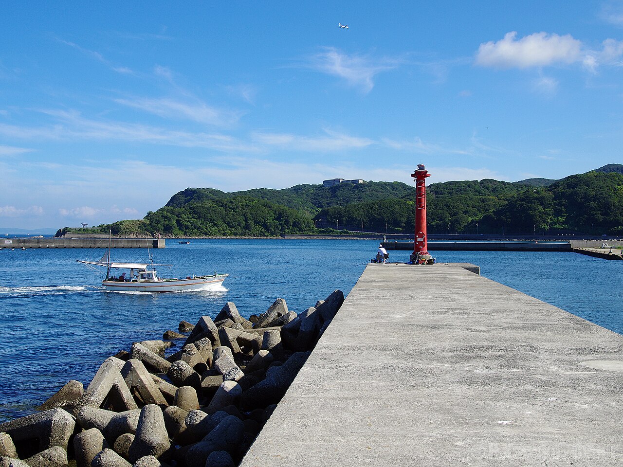 A small boat passing a concrete breakwater with a red beacon at Kada Port, with green hills across the water.