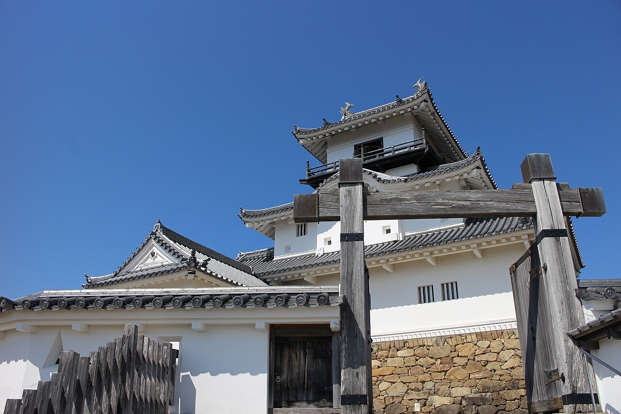 The white three-tier keep of Kakegawa Castle rising behind a weathered wooden gate frame under a clear blue sky