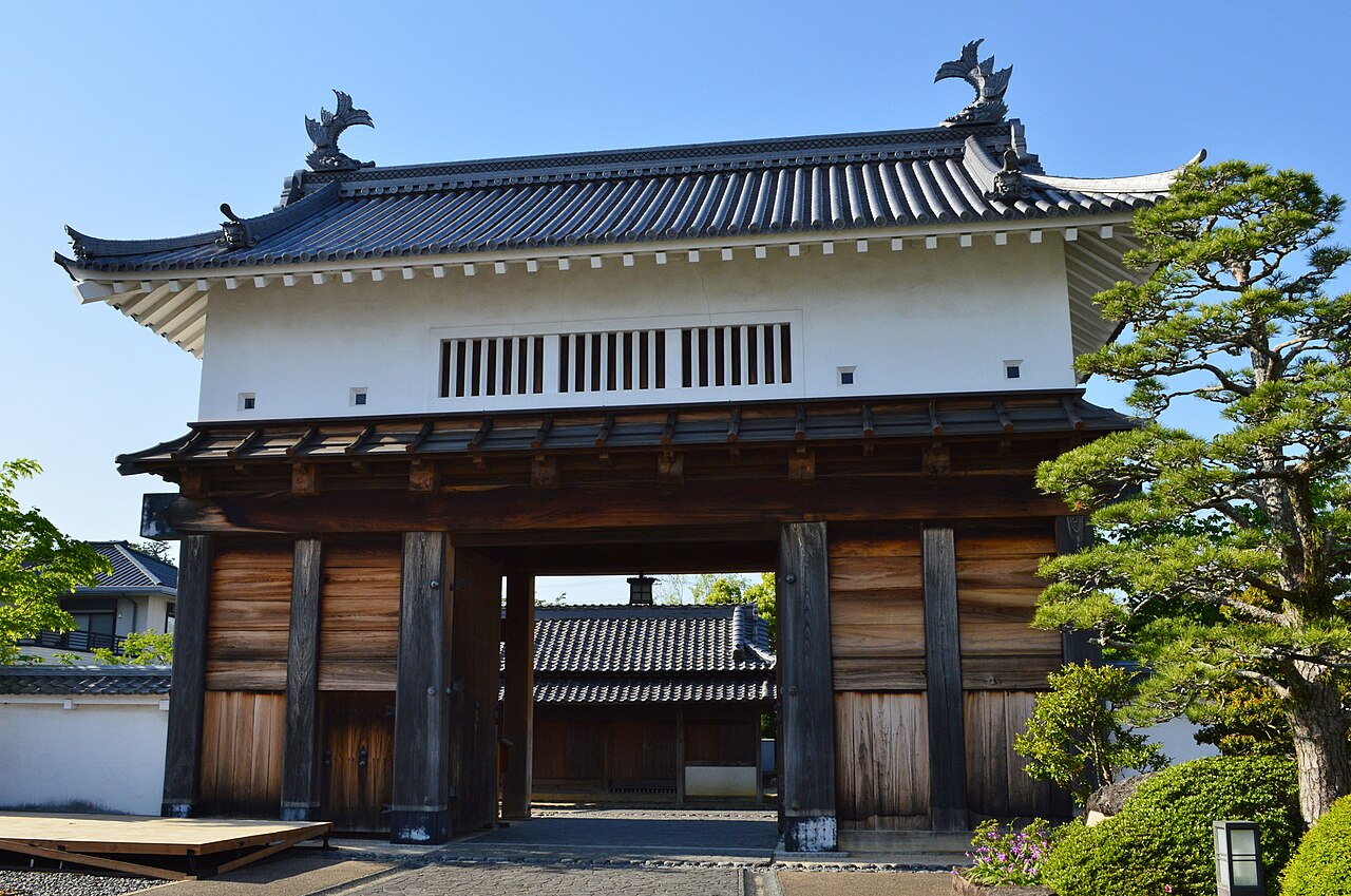 The tiled Otemon gate of Kakegawa Castle with white walls, dark timber posts and a pine tree beside it