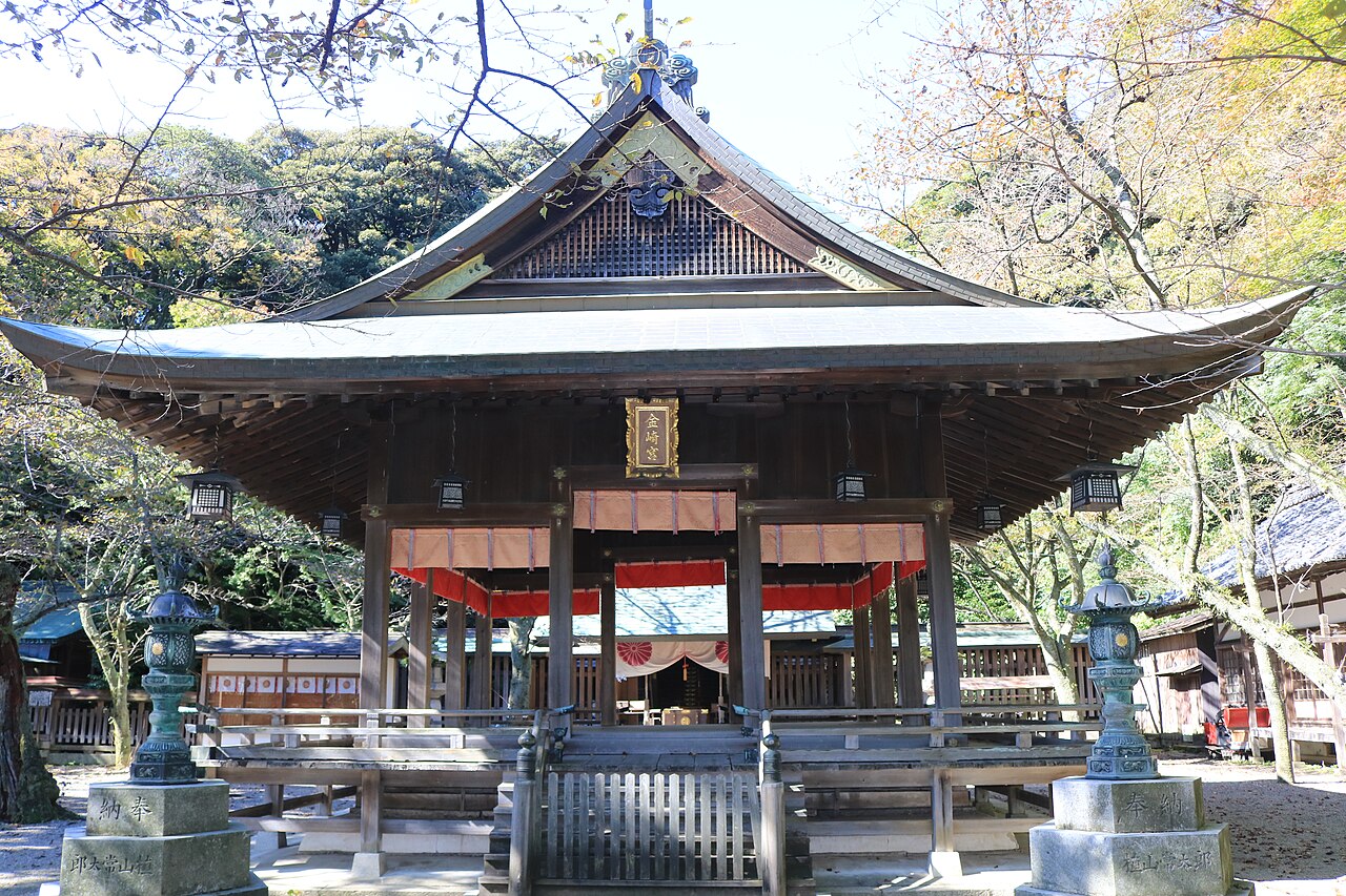 The wooden main hall of Kanegasaki-gu shrine with a curved tiled roof and a hanging nameplate, surrounded by autumn trees.