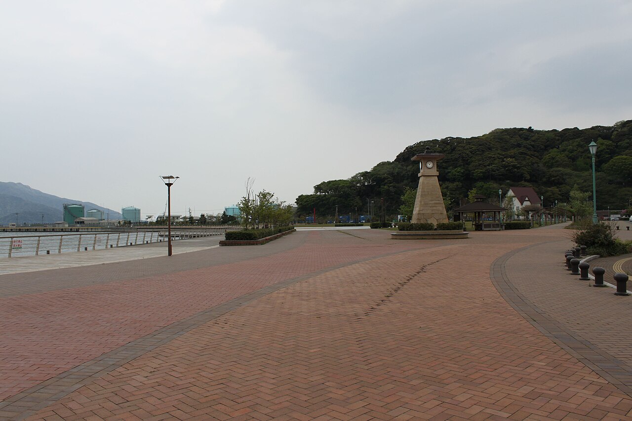 A brick-paved waterfront promenade at Kanegasaki park with a clock-tower monument and wooden benches overlooking the harbor.
