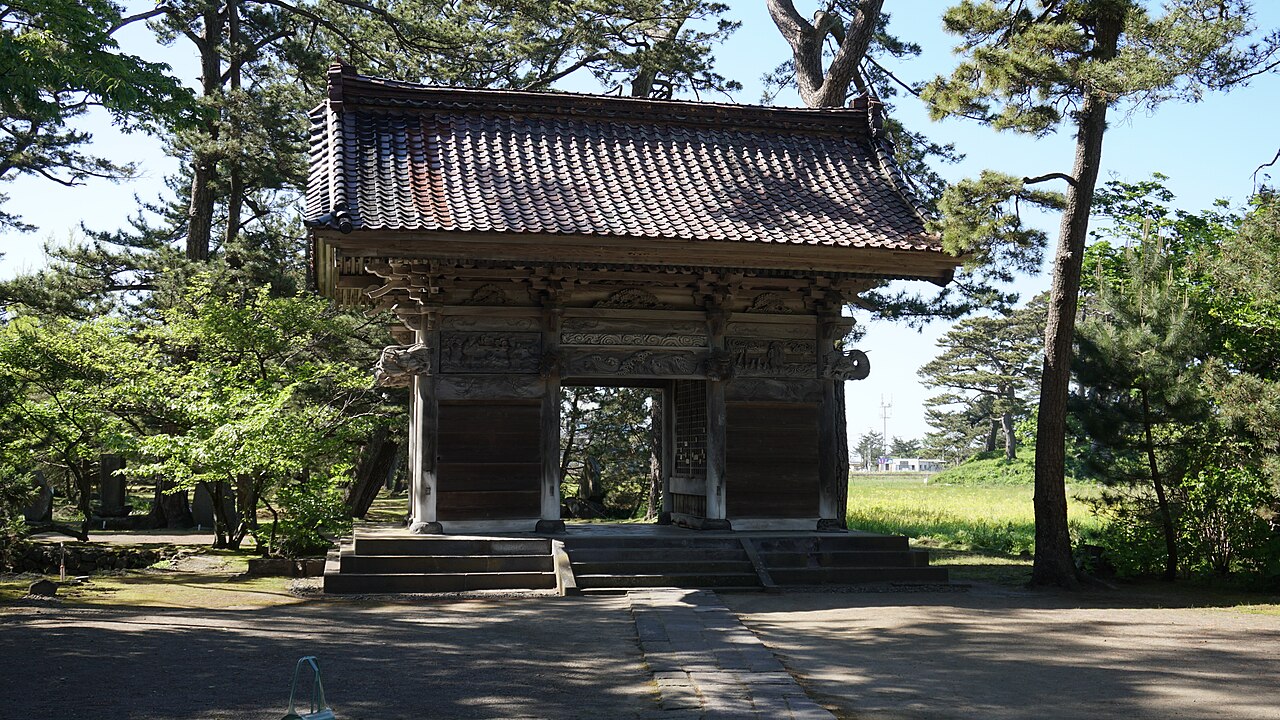 The weathered wooden gate of Kanman-ji temple with a tiled roof and carved panels, framed by pine trees.