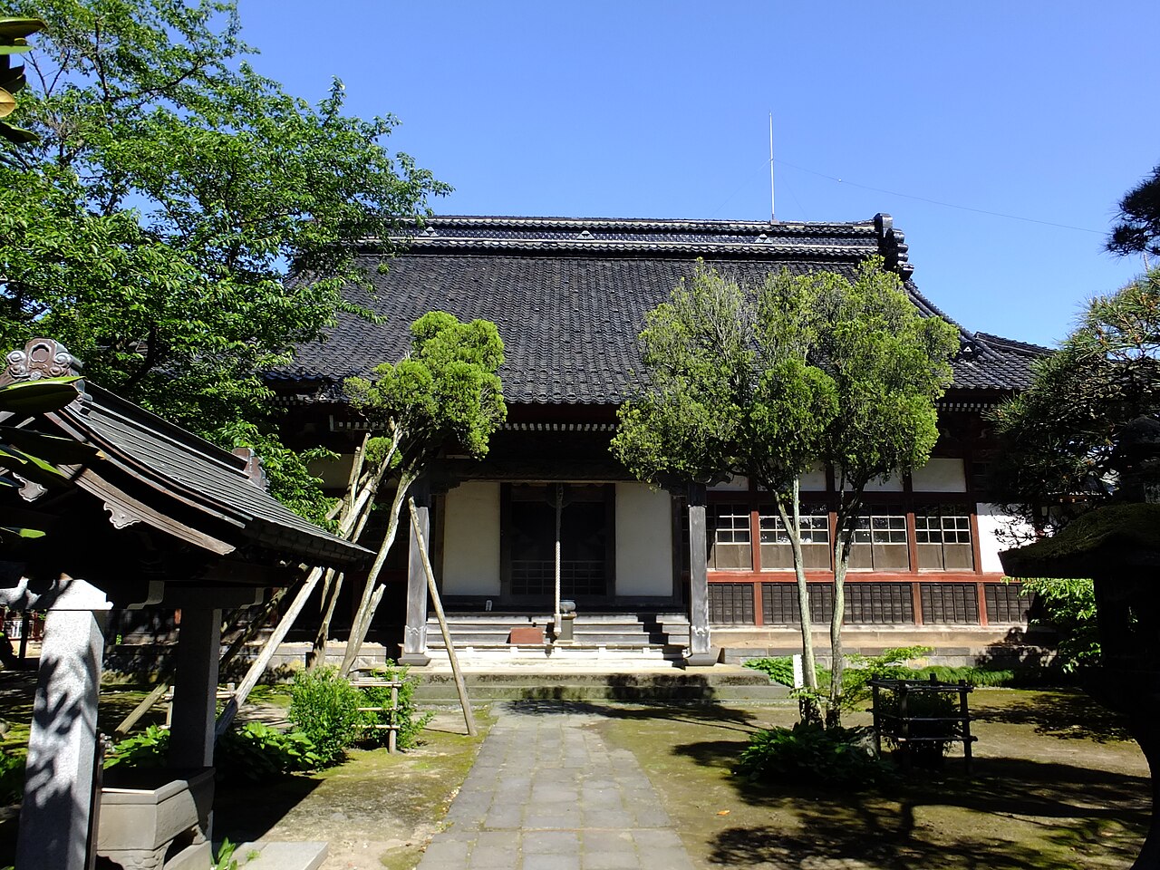 The dark-tiled main hall of Kanman-ji temple seen along a stone path through a small garden of trimmed trees.