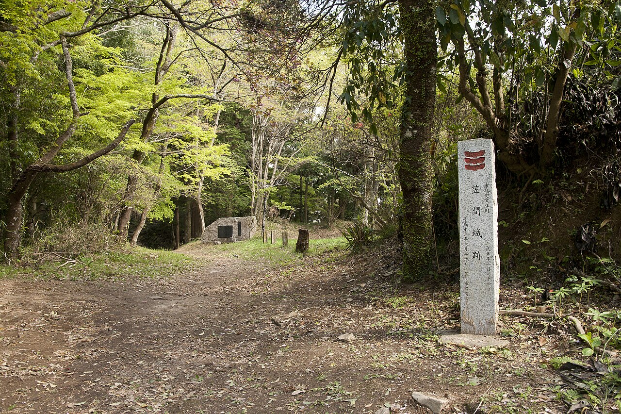 A stone marker reading "笠間城跡" (Kasama Castle ruins) beside a wooded dirt path on Mount Sashiro, with another low stone monument further along the trail.
