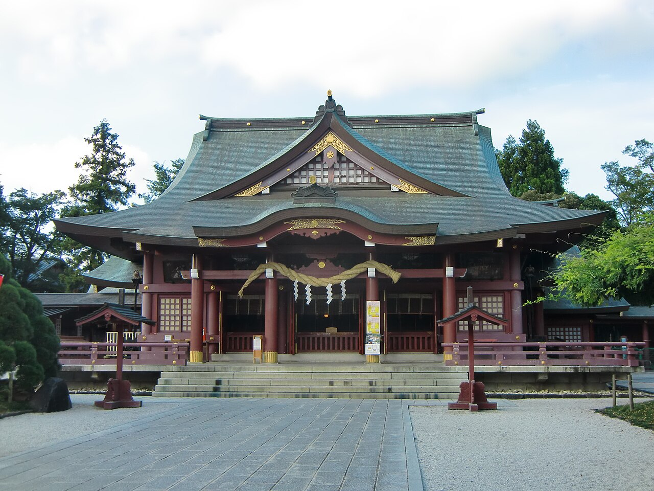 Vermilion-painted main worship hall (haiden) of Kasama Inari Shrine in Kasama, Ibaraki, with a thick shimenawa rope hung across the entrance and stone steps leading up to it.