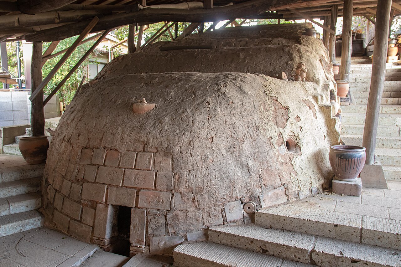 A multi-chambered wood-fired climbing kiln (noborigama) in Kasama under a timber shelter, its clay-covered domes stepping up a slope beside stone stairs and a few glazed pots.
