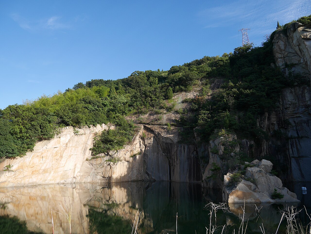 A pale granite quarry wall rising from a still dark pond that mirrors the rock face, with green shrubs along the rim and a clear blue sky above.