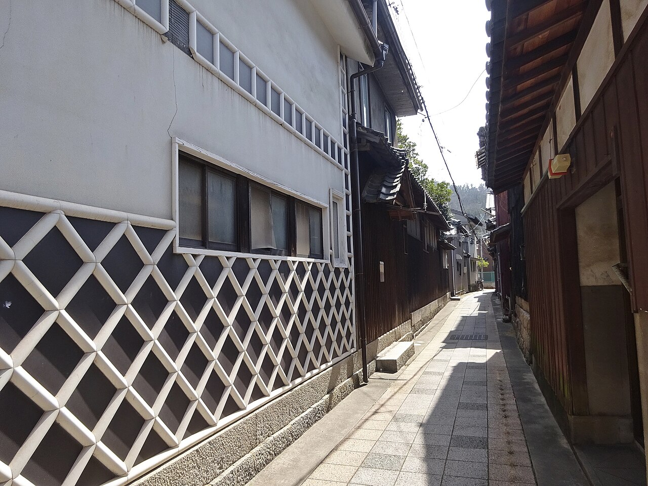 A narrow paved lane between old houses, with a white plaster wall patterned with black diamond lattice on the left, dark wooden walls on the right and a tiled roof ahead, lit by bright sun.