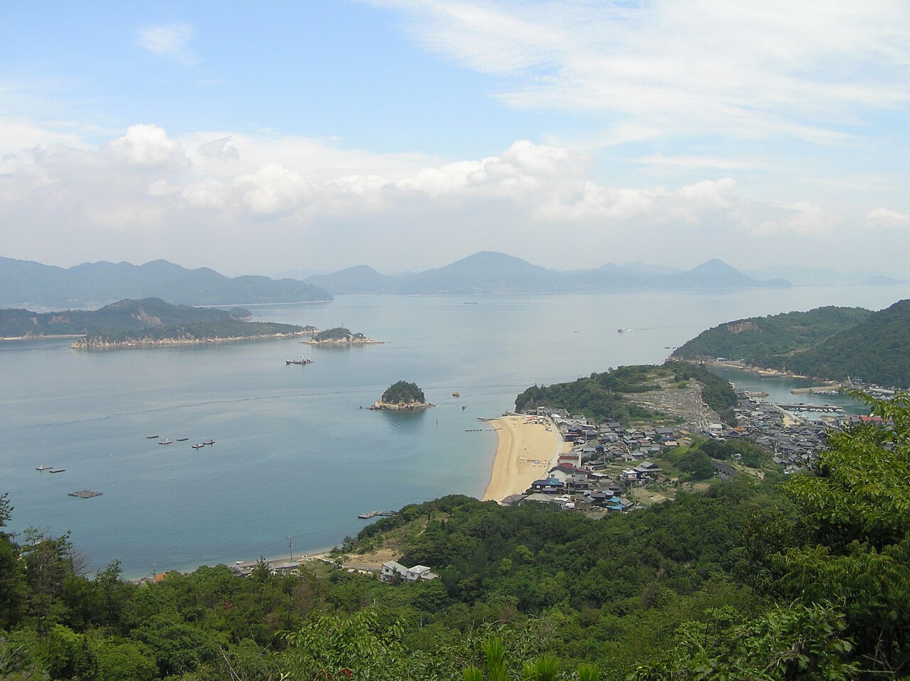 A view from a hillside over a sandy crescent beach and a cluster of tile-roofed houses on a small island, with a small wooded islet just offshore and hazy islands and mountains across a calm blue sea under a partly cloudy sky.