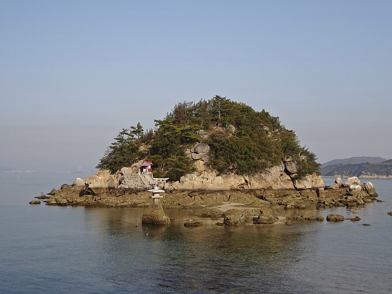 A small rocky islet crowned with pine trees and a tiny red-roofed shrine with a stone torii and lantern, rising from calm shallow water under a pale hazy sky.